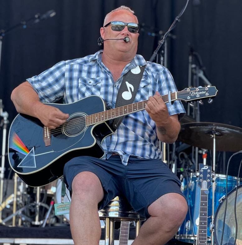 Johnny Sandåker i rutete skjorte spiller på en akustisk gitar med fargerikt design på kroppen, sittende på en krakk foran musikkutstyr.