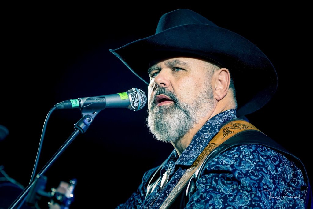 En mann med skjegg, iført cowboyhatt og blåmønstret skjorte, står nær en mikrofon på scenen og opptrer.